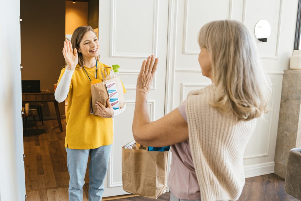 Two women smiling and exchanging grocery bags indoors.